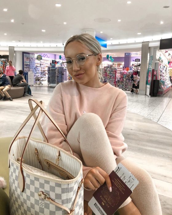 Influencer Elle Darby pide alojamiento gratis a hotel; mujer rubia con lentes de aumento en aeropuerto con VISA en la mano