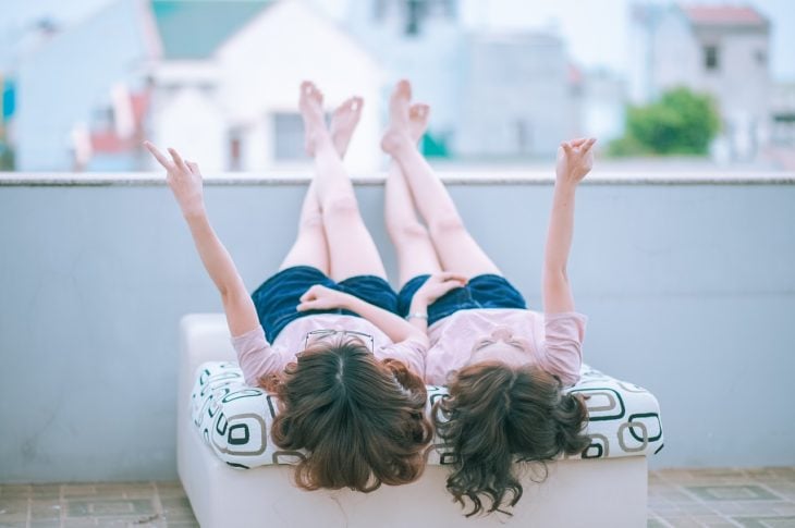dos mujeres acostadas en una cama con playera rosa y short azul con las piernas sobre una barda y levantando un brazo cada una