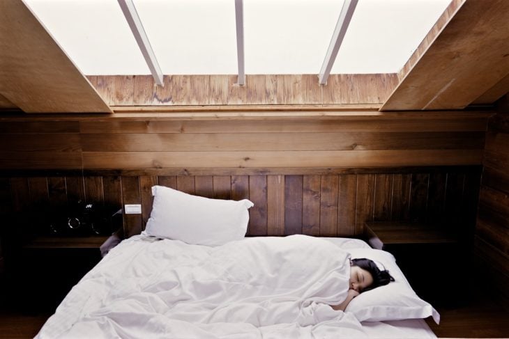 una mujer duerme en una cama con colcha blanca y se observa en la ventana sobre ella la luz del día