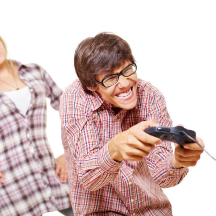un hombre con lentes y camisa cuadrada juega con un control de consola de juegos y atrás se ve el cuerpo de una mujer que lo mira