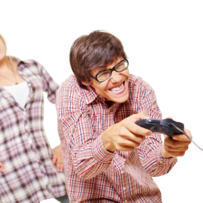 un hombre con lentes y camisa cuadrada juega con un control de consola de juegos y atrás se ve el cuerpo de una mujer que lo mira