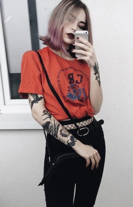 Chica con cabello rosa hasta los hombros, vestida con playera anaranjada y medias de red con jeans; tatuajes en los brazos