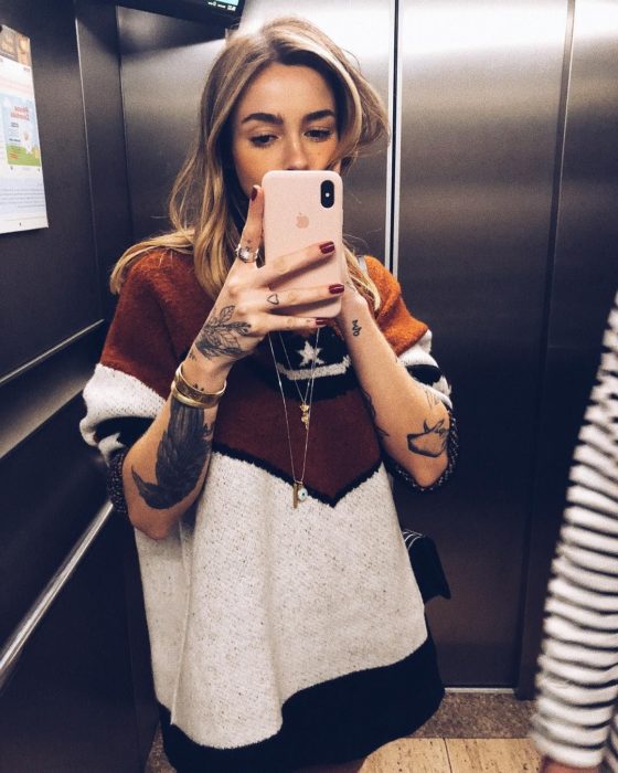 Chica rubia tomándose selfie en espejo del elevador; con suéter tejido oversized, con tatuajes en los brazos y manos