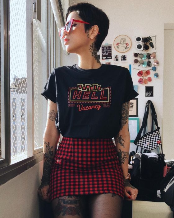 Chica con cabello negro y corte pixie, con lentes retro rojos, playera con estampado y falda de cuadros negros y rojos, look estilo noventero, con tatuajes en los brazos y piernas