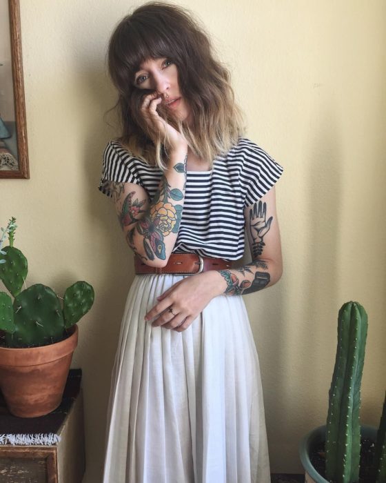 Chica con look estilo boho, cabello a los hombros con degradado ombré, blusa de rayas horizontales, falta blanca y larga, con cinto; tatuajes old school en los brazos; macetas con cactus