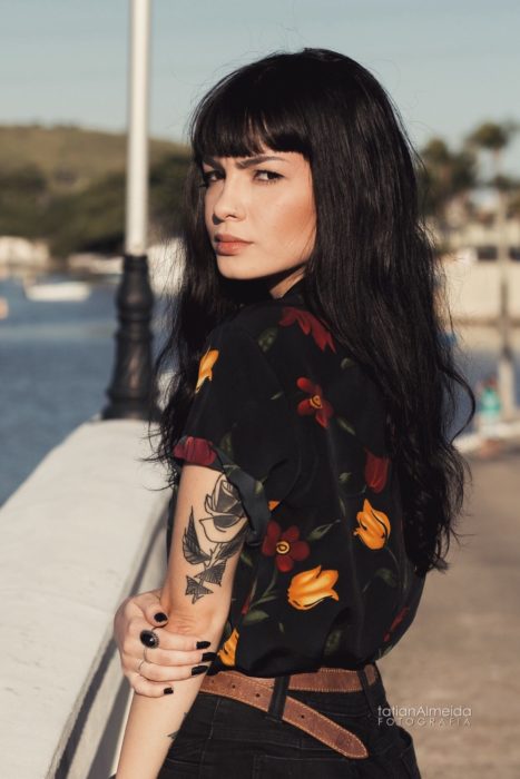 Chica de espaldas, con cabello largo, negro y con fleco, piercing en la nariz y tatuaje en el brazo; camisa negra con flores