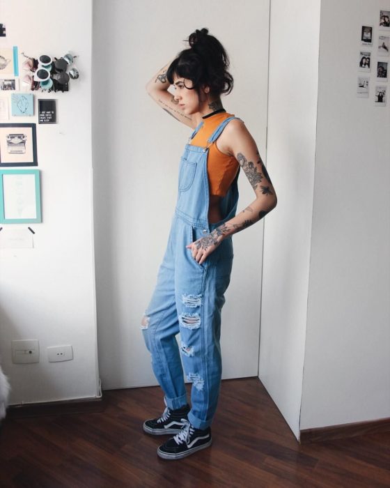 Chica de cabello negro y corto, con overol azul y blusa anaranjada sin mangas, con tatuajes en los brazos y cuello