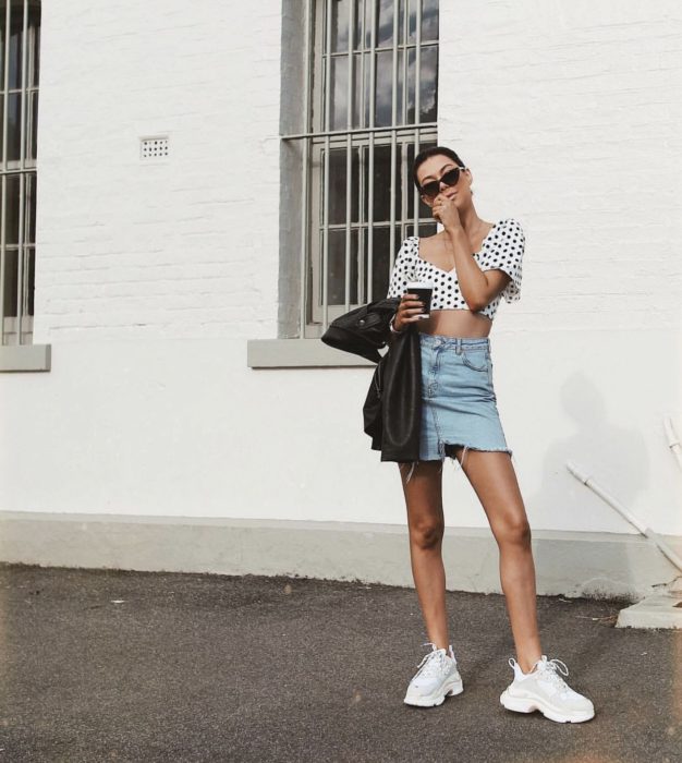 Chica usando una falda de jean, blusa blanca con estampados en negro y unos ugly sneakers de color blanco 