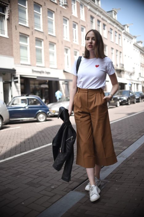 Chica usando unos pantalones estilo culotte con una camisa blanca y tenis, mientras espera parada en la calle