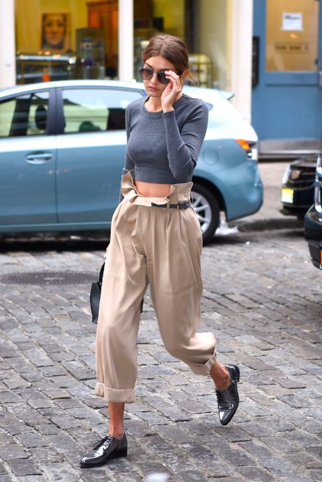 Bella Hadid caminando por la calle mientras usa un pantalón de color beige camisa gris y zapatos de charol