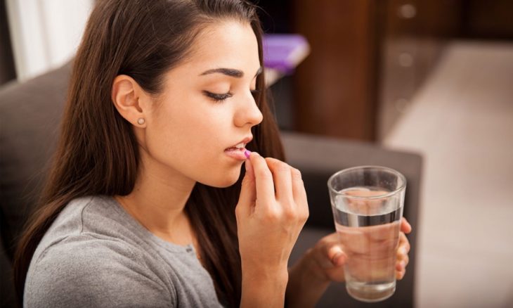 una mujer pone una pastilla en su boca y sostiene con la otra mano un vaso con agua