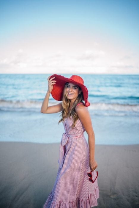 Chic en la playa sosteniendo su sombrero grande y de color rojo 