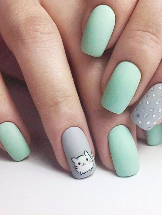 Mano de mujer con uñas pintadas en color verde menta y gris con puntos en tonos claros y un gato en blanco