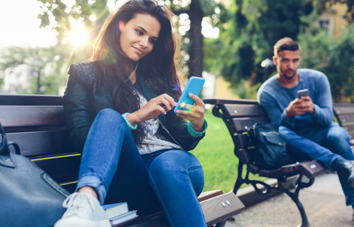 Mujer viendo el celular en un parque observada por hombre con celular