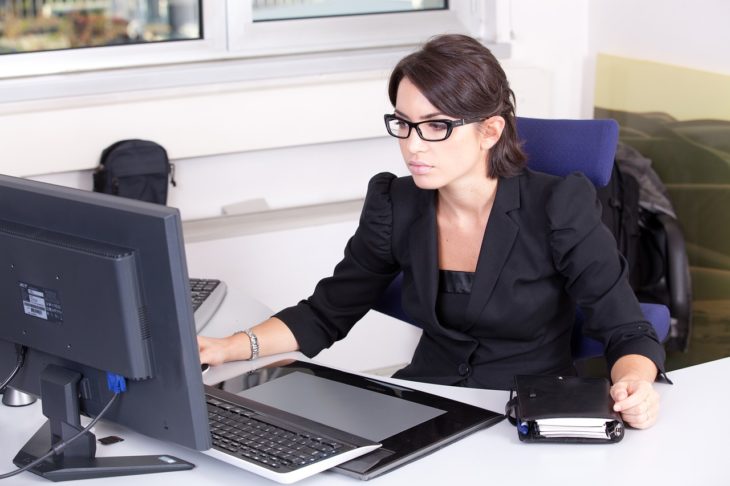 mujer de traje negro y lentes viendo su computadora de escritorio en una oficina