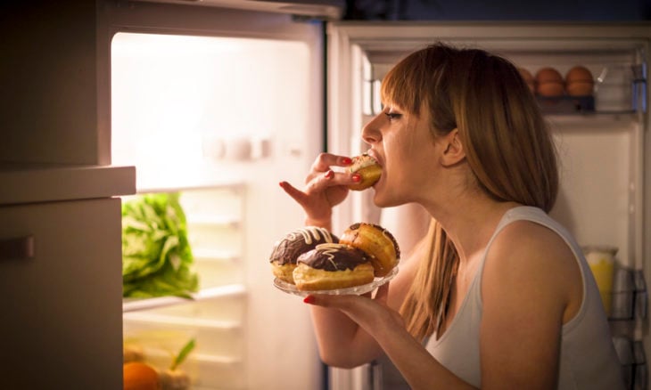 Mujer comiendo panes frente al refrigerador a altas horas de la noche