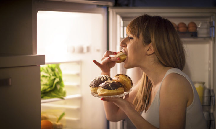 Mujer comiendo panes frente al refrigerador a altas horas de la noche