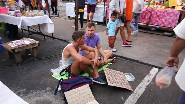 dos jóvenes occidentales sentados en medio de un mercado con carteles pidiendo dinero por que se quedaron sin recursos para viajar