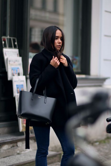 Chica caminando por la calle con un abrigo negro y bolso de color negro 