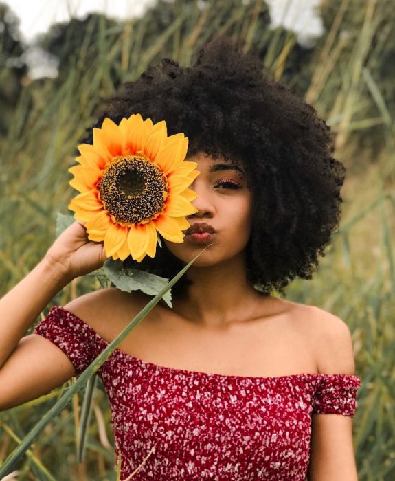 Mujer afroamericana con cabello chino esponjado en un campo con girasol en la mano