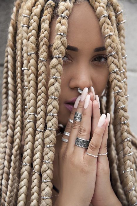 Chica afroamericana con cabello rubio, trenzas y argollas, uñas postizas rosas y varios anillos en los dedos