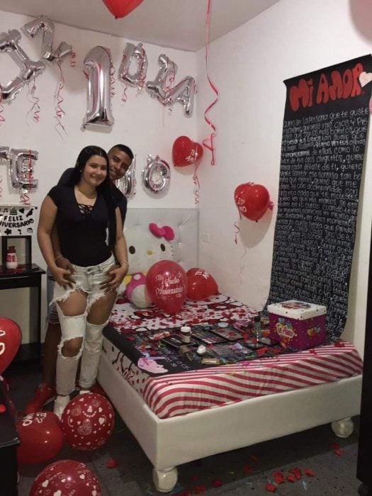Pareja de novios en una habitación decorada con globos, petalos de rosa y pancartas para celebrar su primer día de novios 