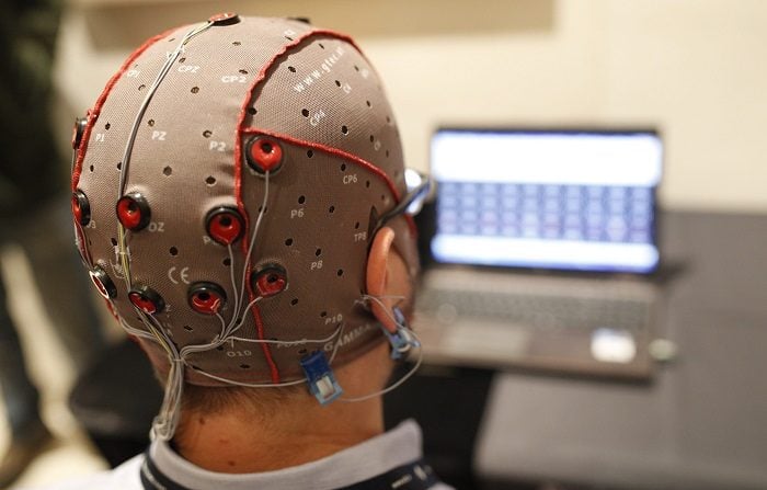 la cabeza de un hombre vista de atrás con una gorra y electrodos conectados y un monitor al frente
