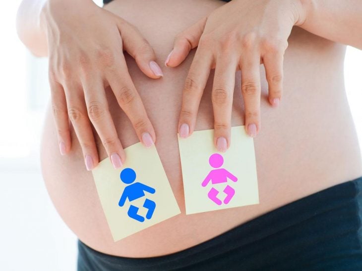 en un vientre de una mujer embarazada sostiene con sus manos dos post-it, uno con una figura en azul y otro con una figura en rosa
