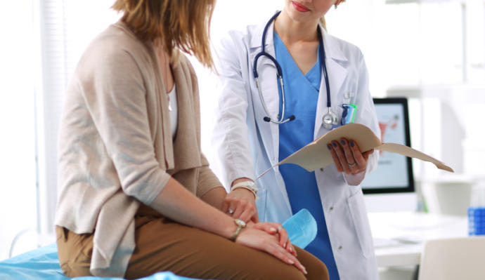 una doctora toma del brazo a una paciente sentada sobre una cama de revisión