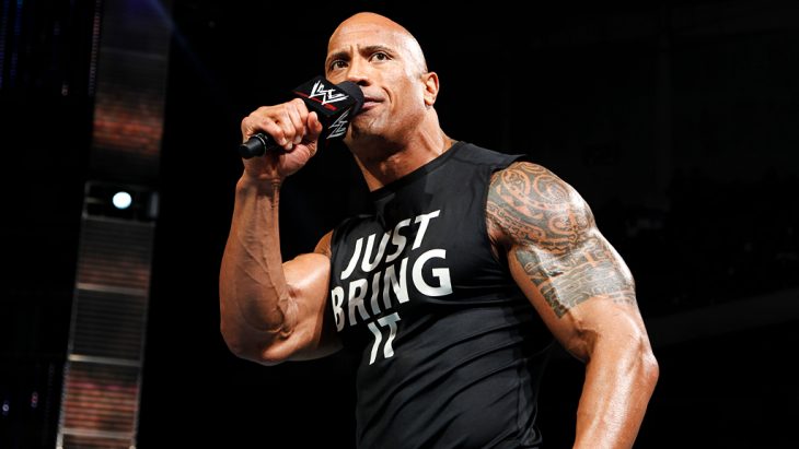 Dwayne Jonhson en camiseta negra con letras blancas y sosteniendo un micrófono de la WWE