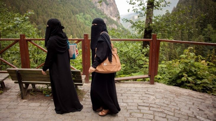 dos mujeres árabes caminan por un espacio abierto en un parque, se observa flora y una montaña