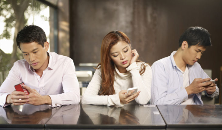 dos hombres y una mujer con rasgos asiáticos en una misma mesa revisando cada uno sus celulares