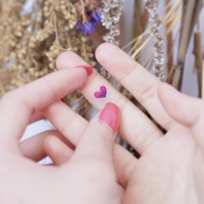 Chinese tattoo artist, Mini Lau;  Small, feminine tattoo with pastel heart colors on the middle finger