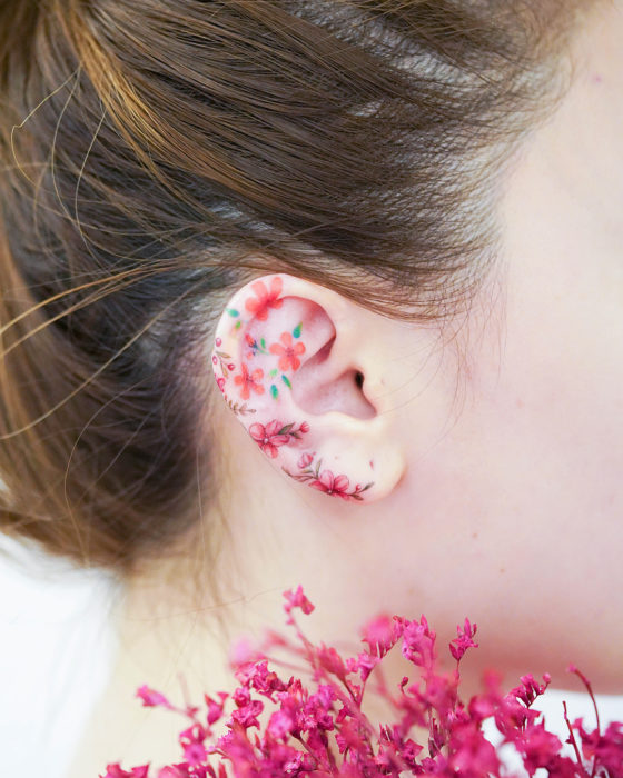 Chinese tattoo artist, Mini Lau;  Small and feminine tattoo with pastel colors of red and orange flowers on the ear