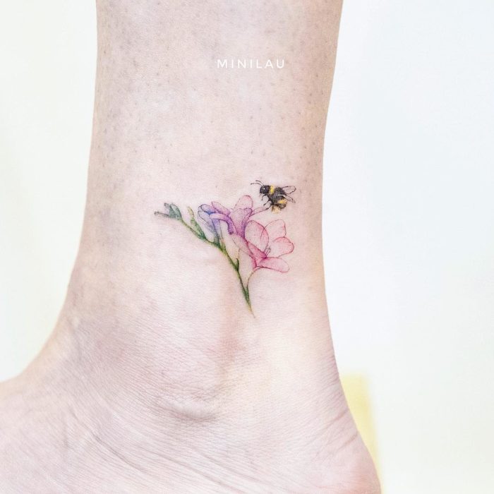 Tatuadora china, Mini Lau; tatuaje pequeño y femenino con colores pastel de abeja en una flor rosa en el tobillo