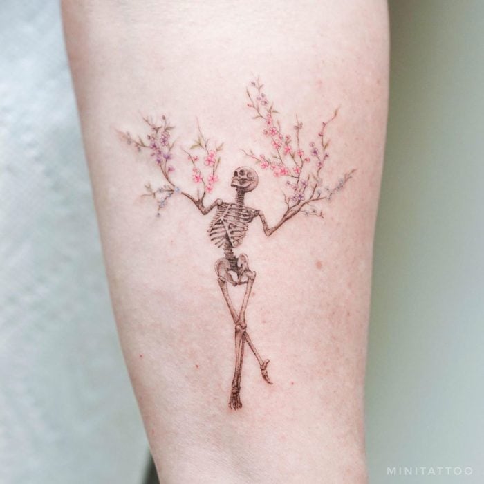 Tatuadora china, Mini Lau; tatuaje pequeño y femenino con colores pastel de esqueleto con brazos de ramas y flores