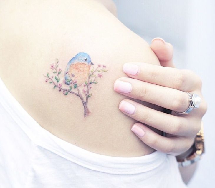 Tatuadora china, Mini Lau; tatuaje pequeño y femenino con colores pastel de pájaro azul en una rama con flores rosas