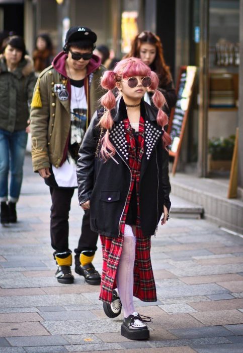 Moda japonesa harajuku; niña de cabello rosa con peinado de trenzas, con vestido de estampado esconcés y chamarra de piel oversized con estoperoles y zapatos de plataforma