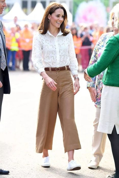 Kate Middleton usando unos pantalones culottes de color café, blusa blanca y tenis de color blanco 