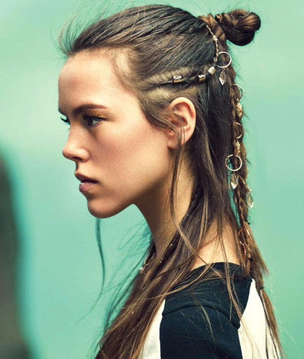 Chica de cabello castaño y lacio peinada como Lagertha de Vikingos, con trenzas, argollas y un medio chongo