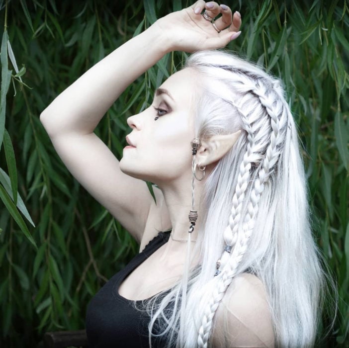Chica muy pálida, con orejas de elfo, de cabello largo y blanco con peinado estilo élfico o vikingo con trenzas de lado y argollas