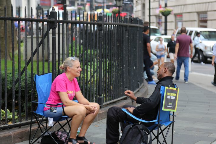 Una voluntaria de Sidewalk talk sentada en una silla colocada en una banqueta escucha a un joven sentado frente a ella