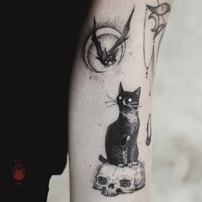 Tatuaje de halloween, gato negro sobre un cráneo mirando a un murciélago en la luna