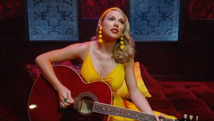 Escena del video Lover de Taylor Swift, está tocando la guitarra sentada en un sillón rojo oscuro y con vestido amarillo y aretes amarillos largos