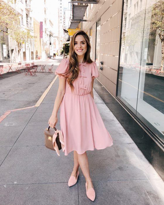 Chica sonriendo en la calle, con cabello castaño peinado hacia un lado, con vestido rosa casual