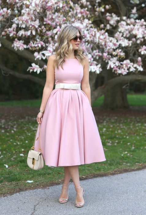 Chica rubia con lentes de sol y vestido rosa liso sin mangas, frente a un árbol floreado