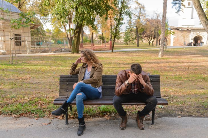 Hombre agarrándose la cara y mujer volteada, ambos sentados en la banca de un parque