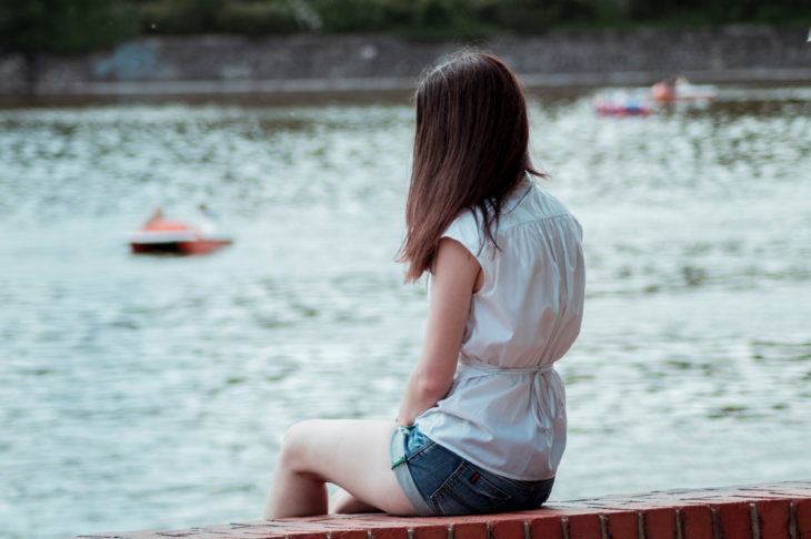 una mujer sentada a la orilla de un lago, sola, con blusa blanca, de espaldas