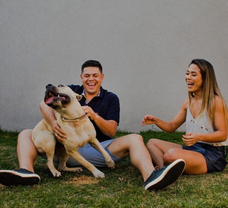 Pareja de novios sentados en el pasto mientras rien y juegan con su perro