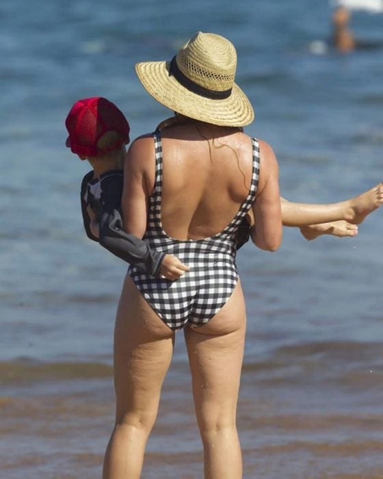 Foto de espaldas de Hilary Duff con su hijo en brazos frente a una playa, usa un traje de baño de cuadros y sombrero